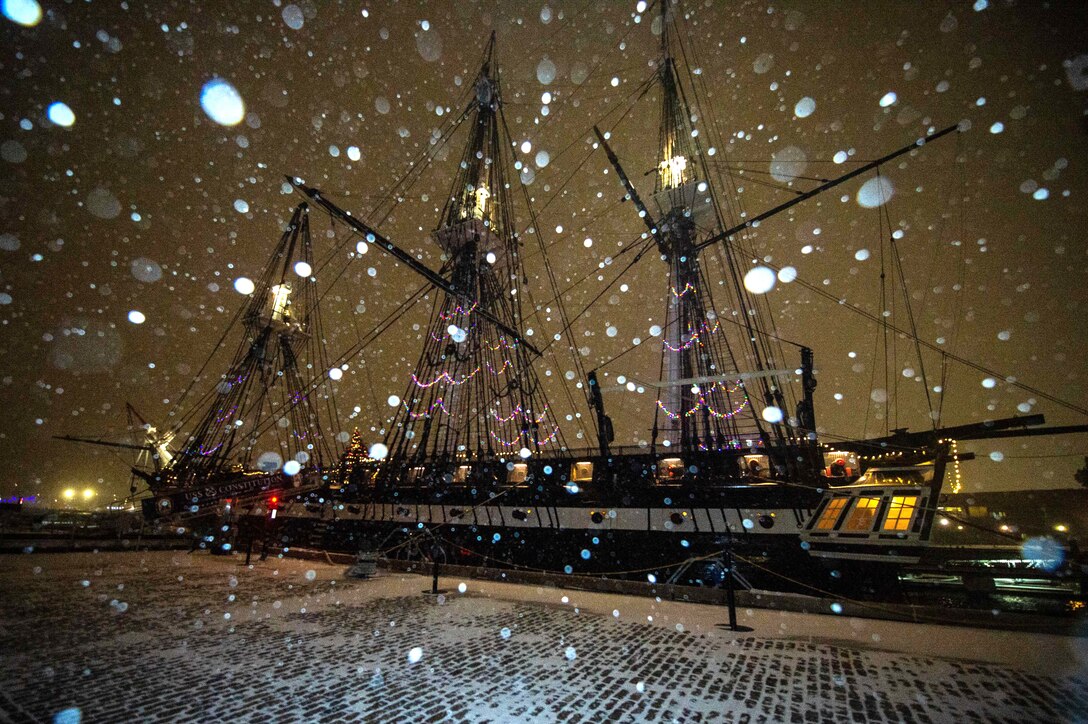 A moored three-masted ship displays holiday lights and decorations as snow falls around it at night.