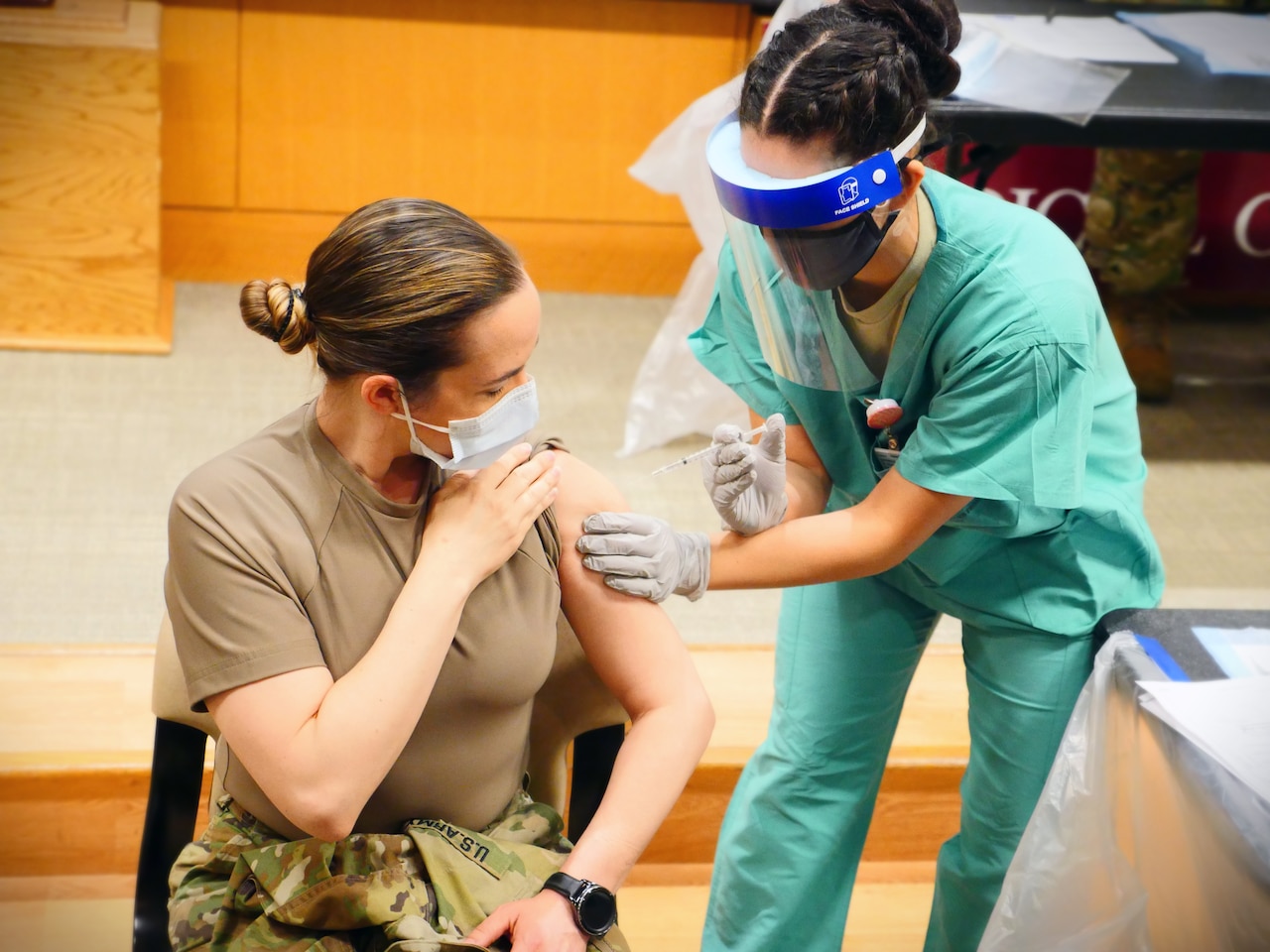 An airman wearing protective gear gives a soldier wearing a face mask a shot.