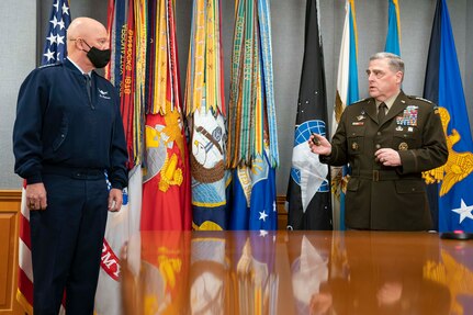 Two men separated by a table talk to one another; behind them is a row of flags.