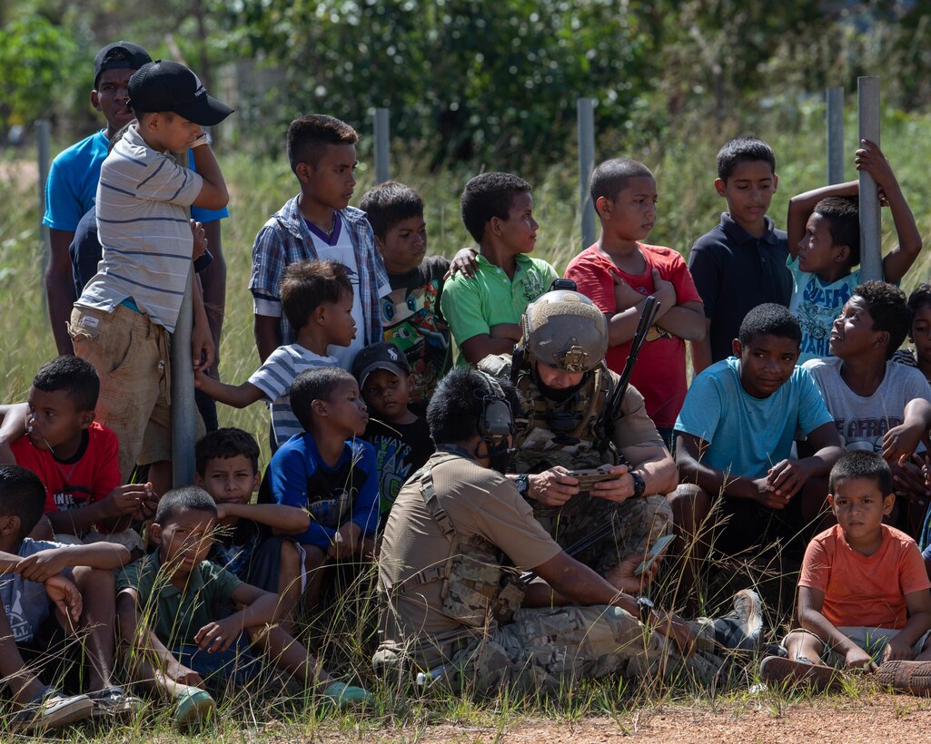 Honduran children in shorts and tee-shirts huddle around two Special Tactics Operators, one sitting on the ground, the other squatting and tapping into a tablet.