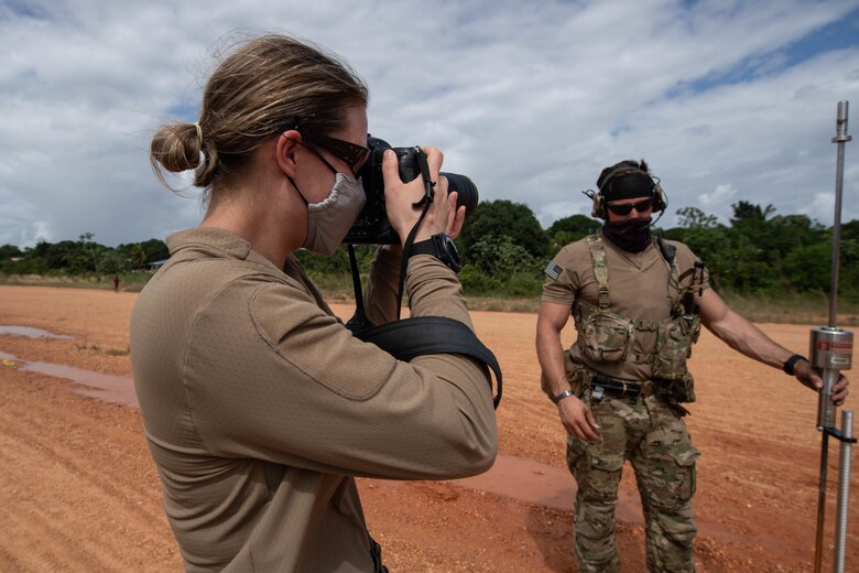 An Air Force combat photographer is show with camera held to her eye photographing a scene off-frame to the right. A Special Tactics operator stands facing us pointing off to his left at the scene being photographed.