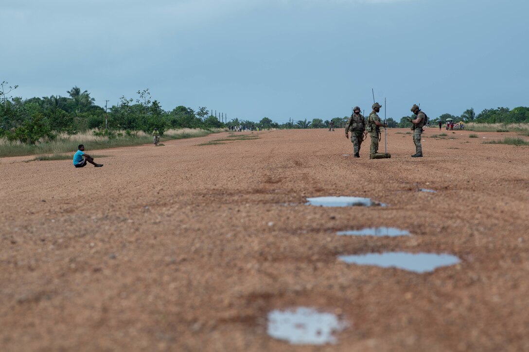 A trio of standing Special Tactics operators gather around a pole in the ground being used to test the hardness of the ground for this potential runway. Two operators are working with the pole while a third walks toward the pair. A young boy has sat down on the ground a ways off to the left of the three operators to watch them work.
