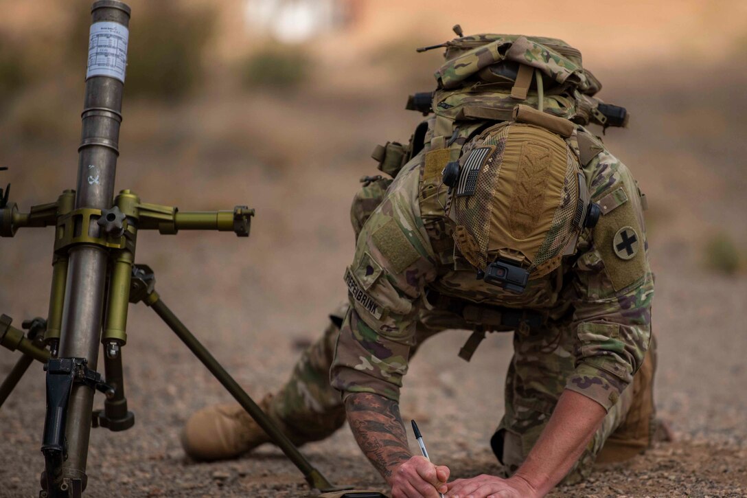 A soldier kneels on the ground to write in a notebook.