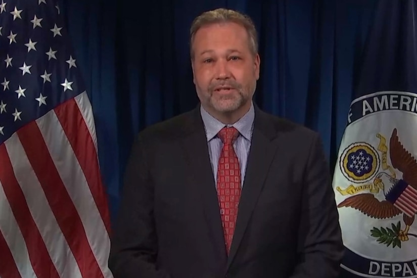 A man looks forward; a flag and a seal are behind him.