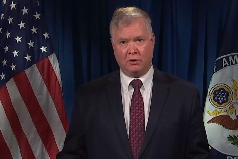 A man flanked by a flag and a seal looks ahead and speaks.