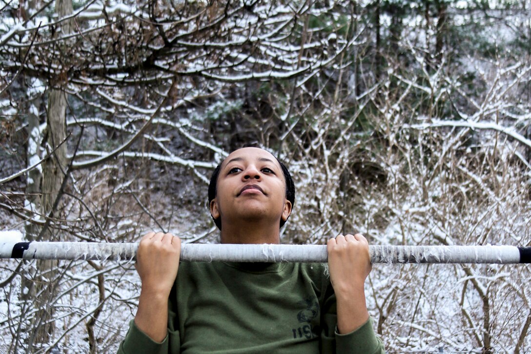 A Marine does a pullup outside, with snow coated trees in the background.