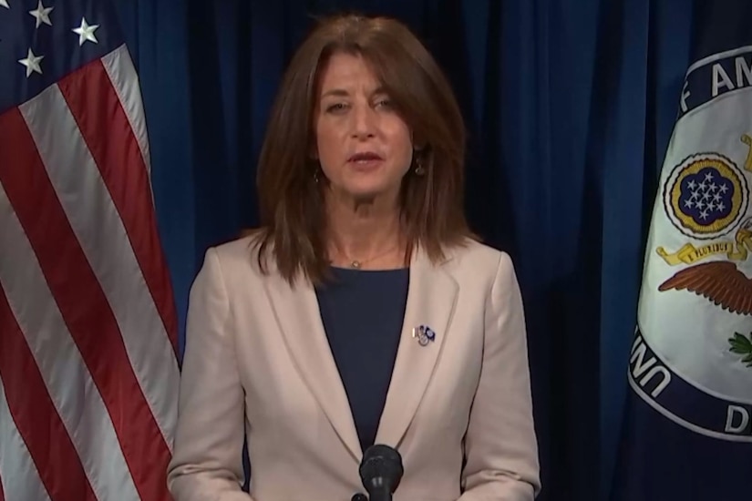 A woman flanked by a flag and a seal looks forward.