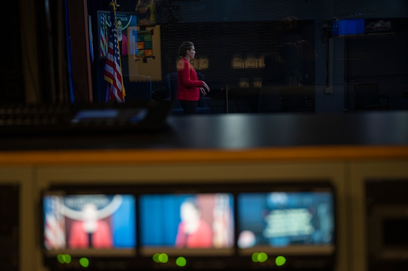 A  woman stands in the background of a row of three video monitors.