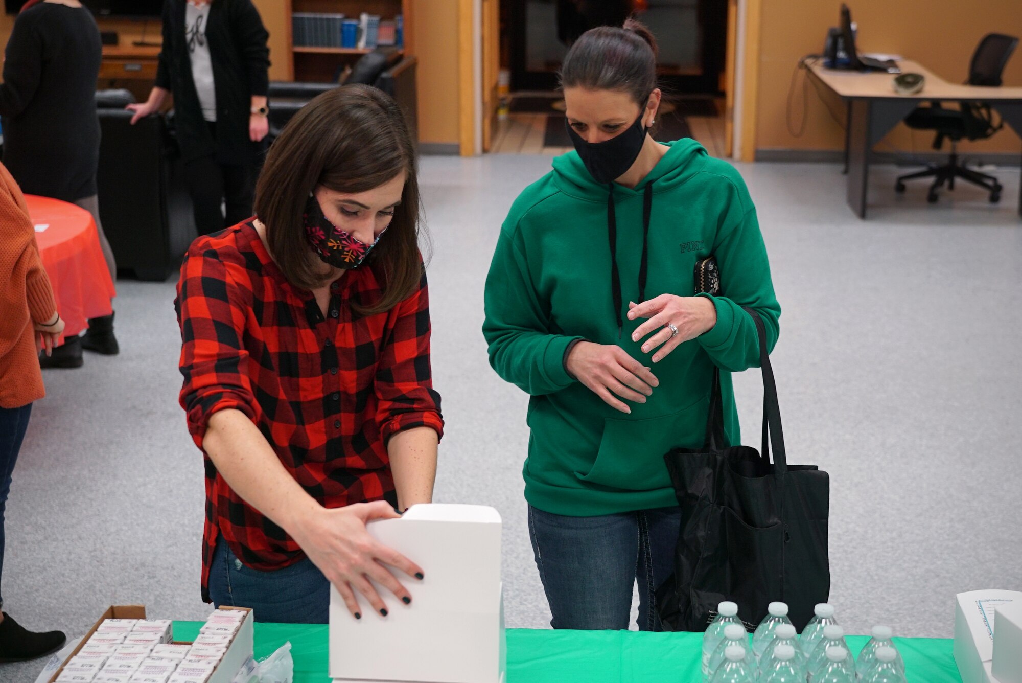 Two women stand center-frame at a table with juice boxes, water bottles and cardboard lunch boxes on top of it.