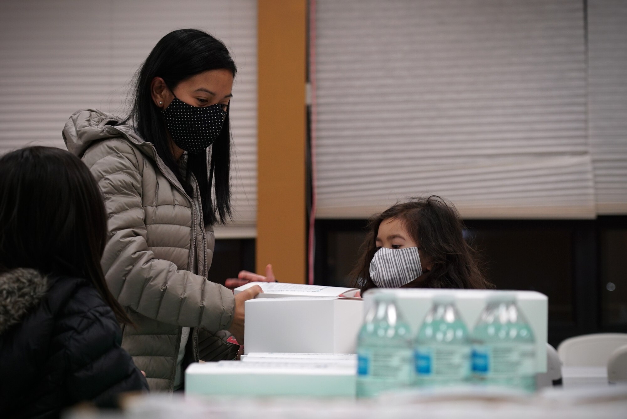 A mother stand on the left side of the frame as her child stands in front of her center-frame, both reaching for a small white cardboard lunch box.