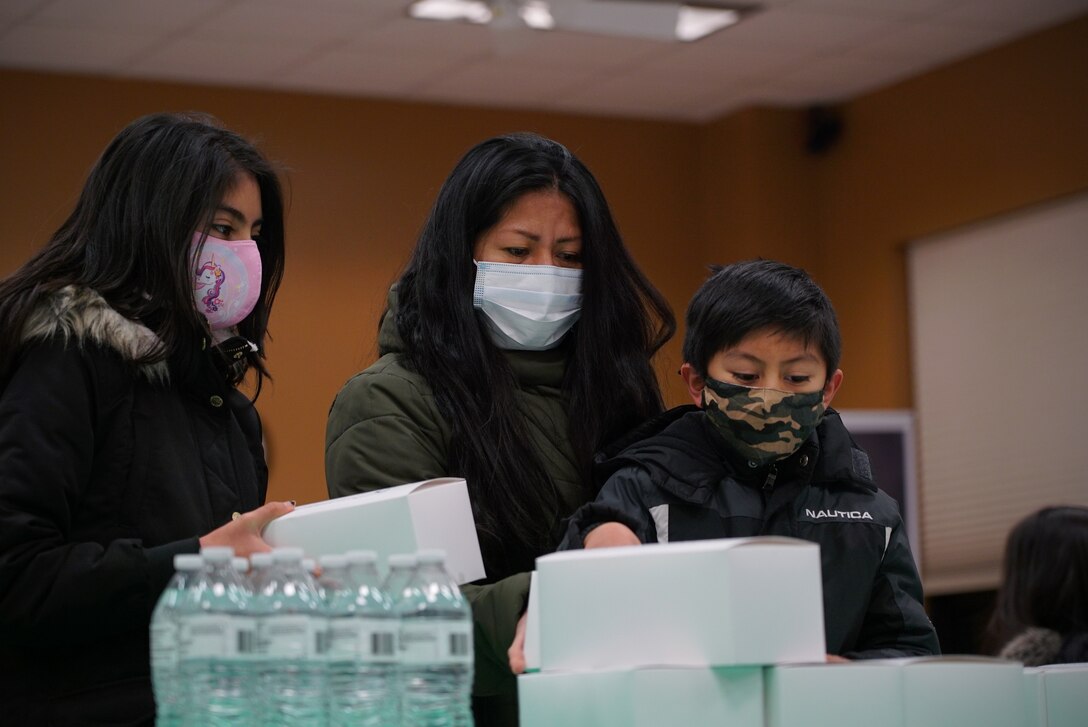 A mother is shown standing between her two children in the background, picking up white meal boxes in the foreground.