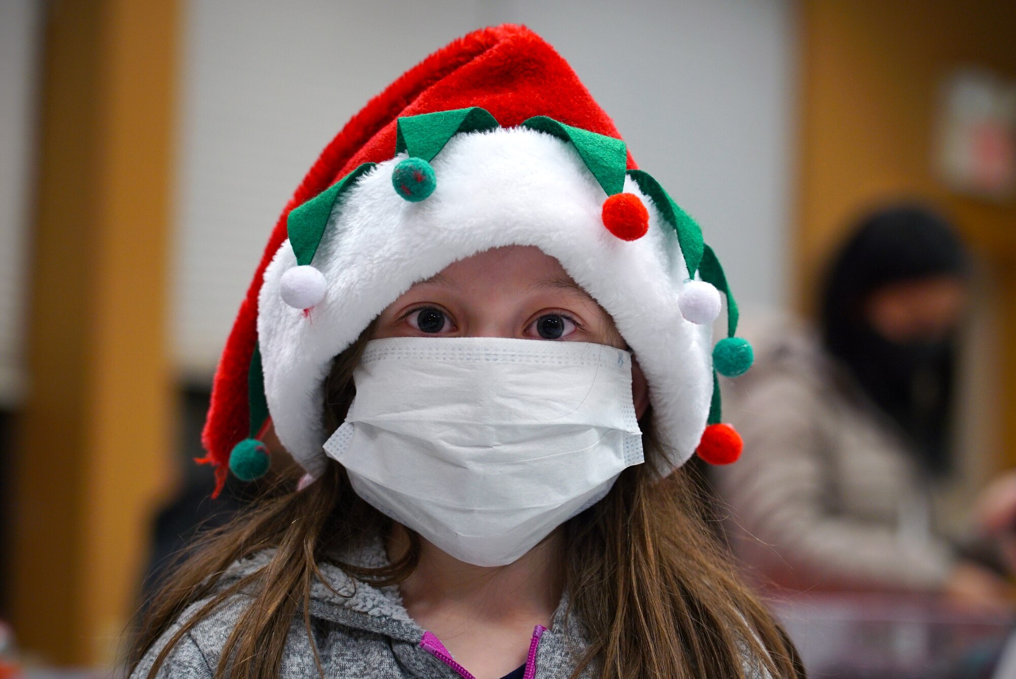 A young girl is shown center-frame wearing a Santa hat and medical face mask stares into the camera.