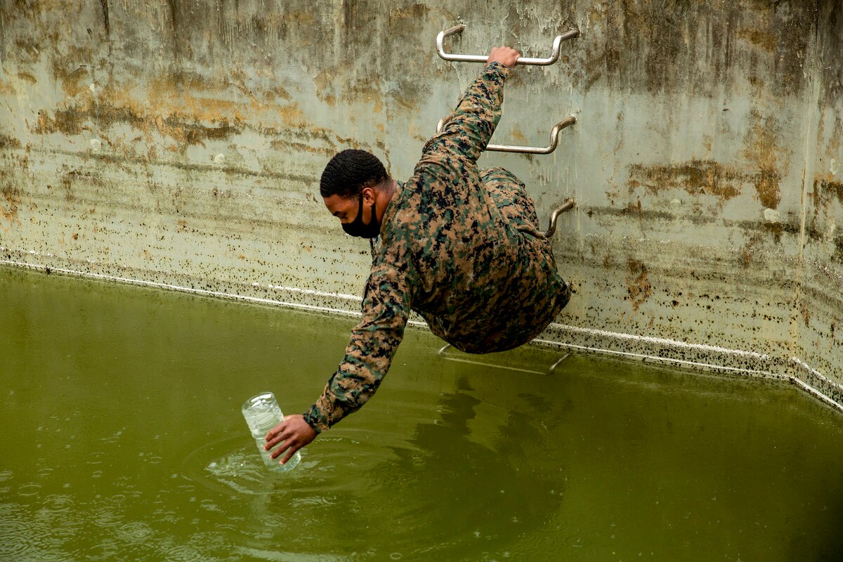 A Marine holds onto a ladder with one hand and reaches a cup into a pool of green water beneath him with the other.
