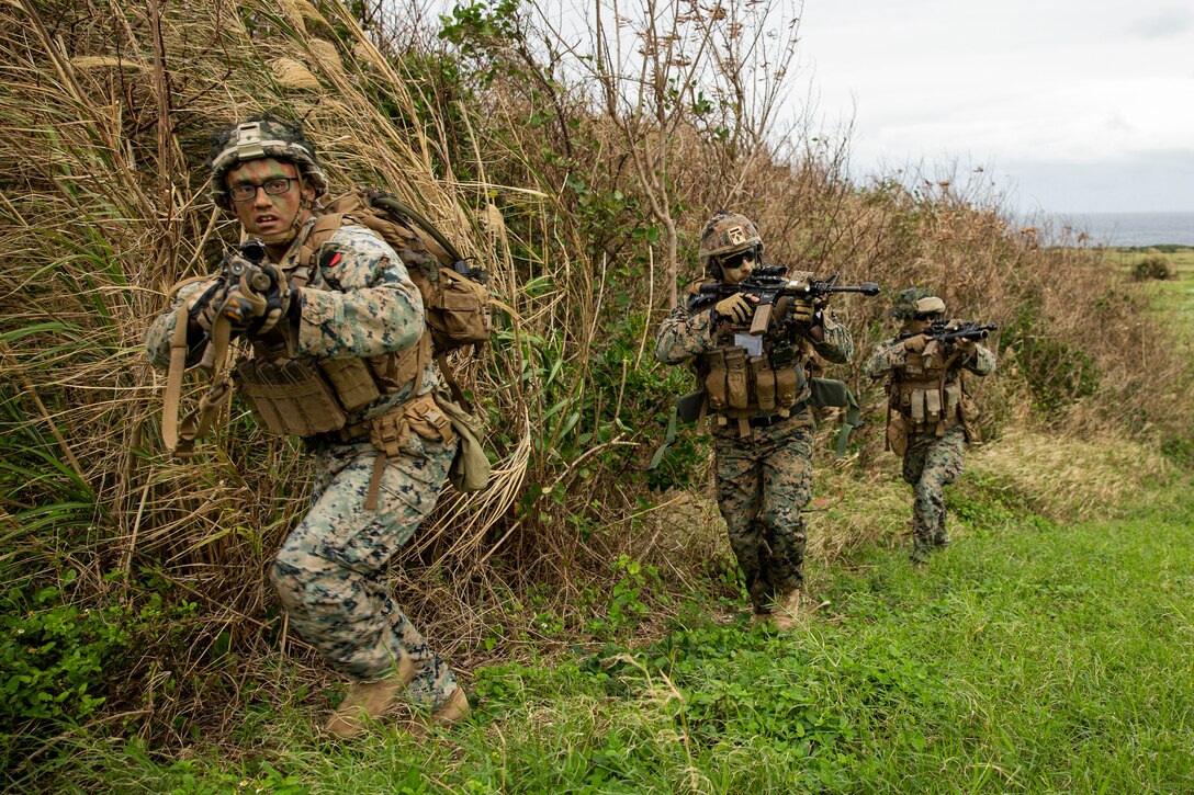 Three Marines holding weapons walk along the edge of a field.