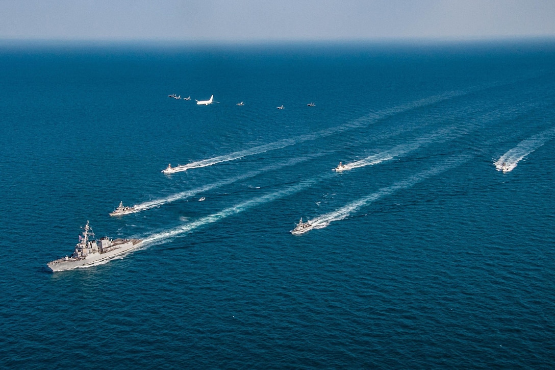 A large group of ships steam across the open water with aircraft flying above them.