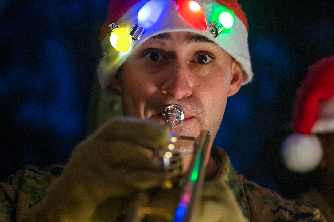 A Marine wearing a Santa hat with Christmas lights on it plays a trumpet.