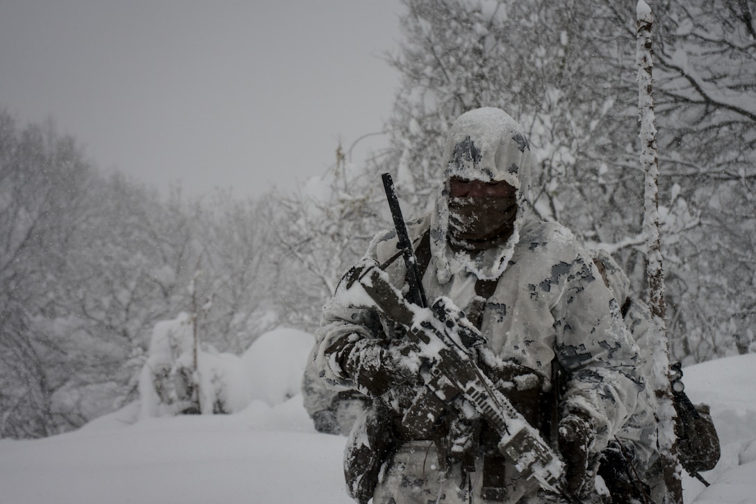 A Marine covered in snow walks through the woods carrying a weapon.