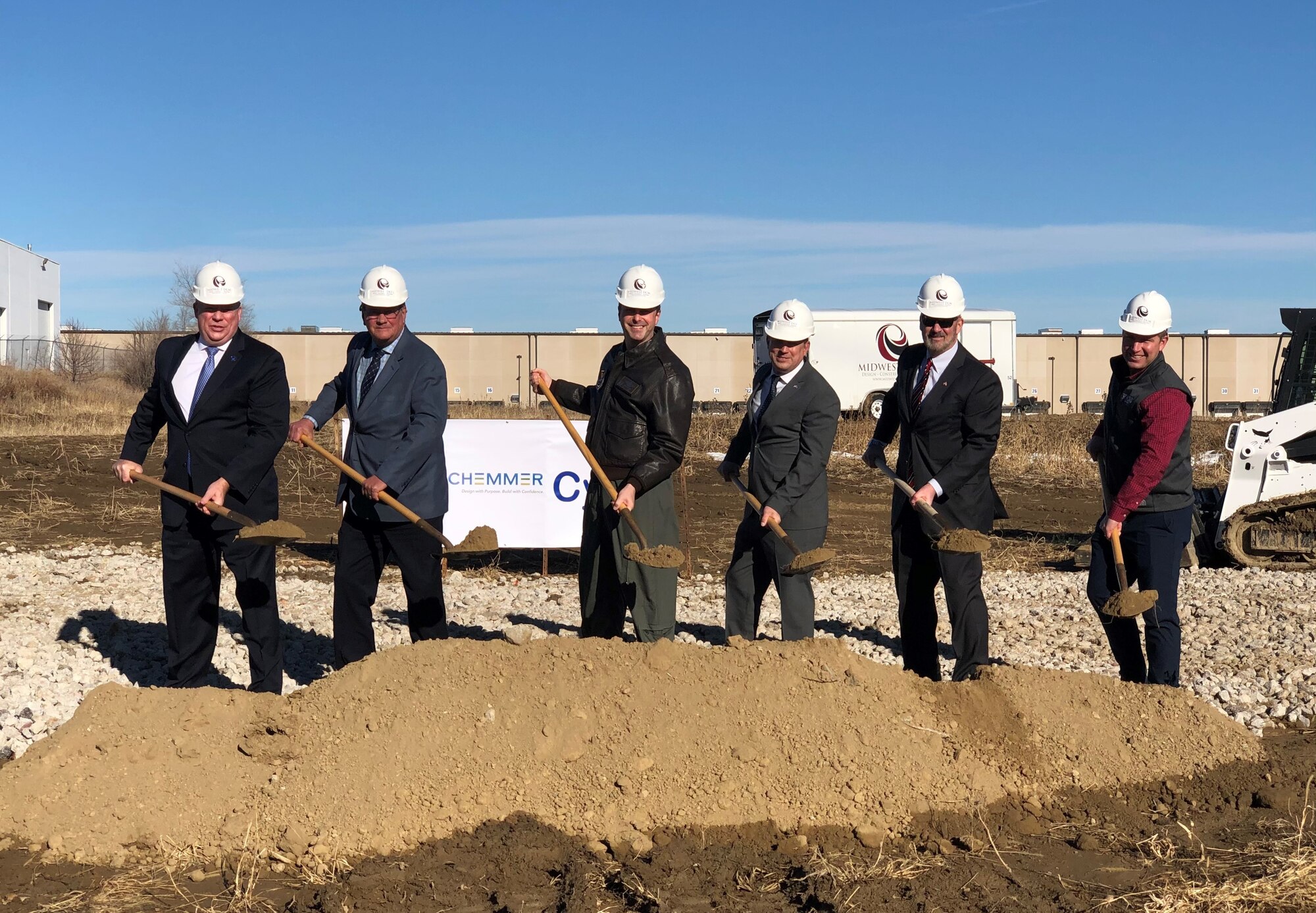 Six men stand in a line in front of a pile of dirt with shovels at a ground-breaking ceremony