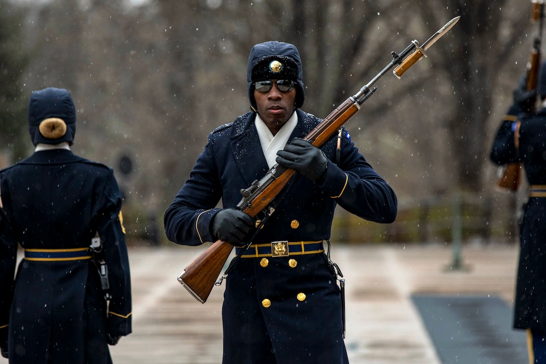 Three soldiers stand or walk on pavement as snow falls around them.