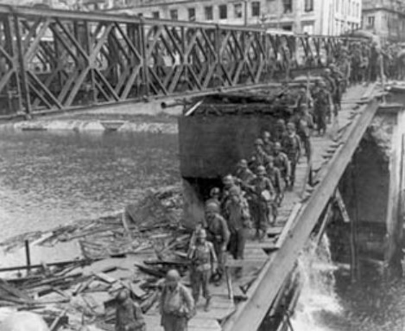 Dozens of soldiers march across a foot bridge over a river.