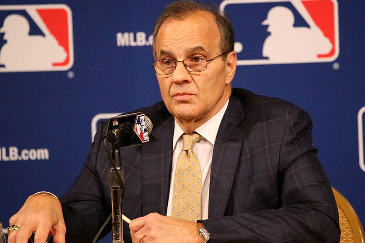 A man, seated at a microphone, speaks. The Major League Baseball logo and URL are in the background.