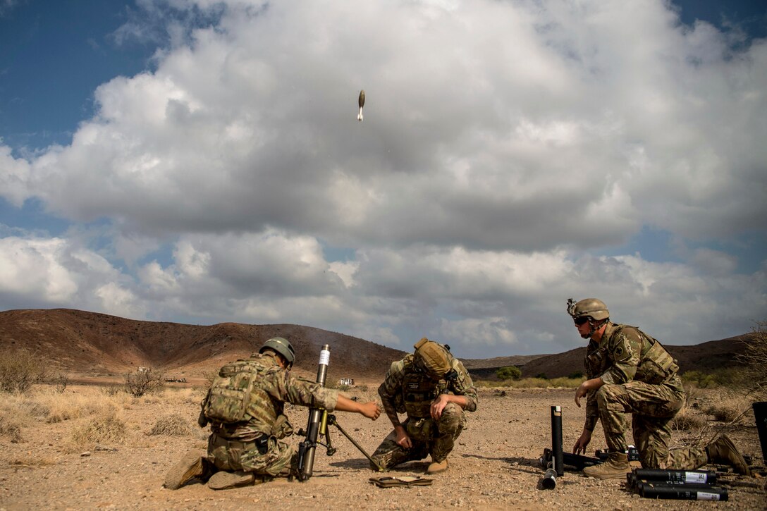 Three soldiers kneel or crouch while firing a mortar system in desert-like terrain.