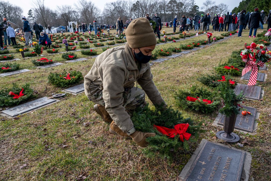 An airman kneels and places a wreath with a red ribbon at a gravesite in a cemetery where other people gather to do the same.