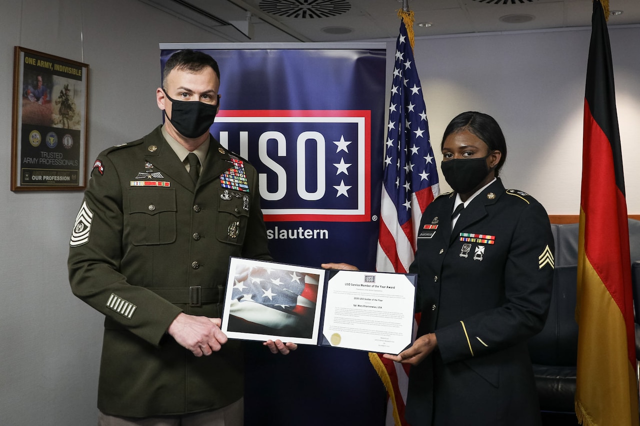 Two service members in masks hold up an award certificate in front of a USO sign.