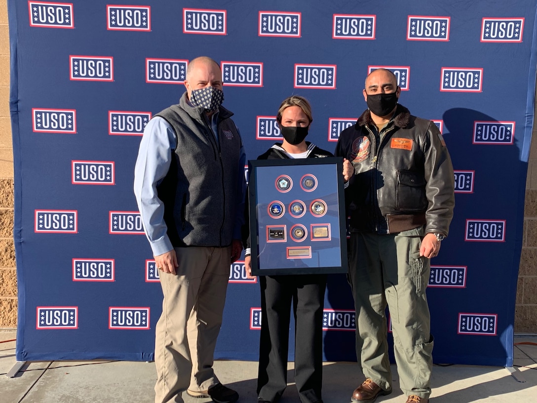 A sailor standing between two men holds a plaque. A USO backdrop is behind them.