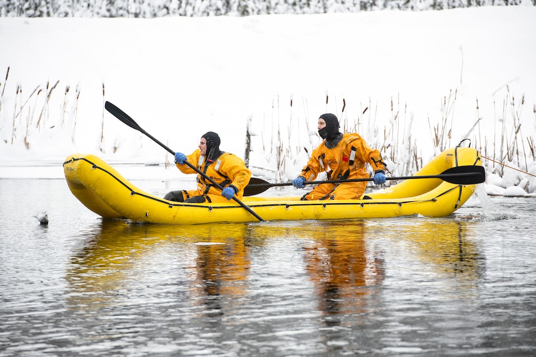 Two airmen in yellow protective gear paddle a yellow inflatable boat in a lake surrounded by snow.