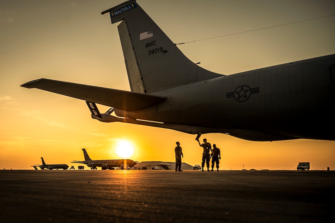 Airmen, shown in silhouette, stand beneath an aircraft on a flightline, with a low sun and gold-orange sky in the background.