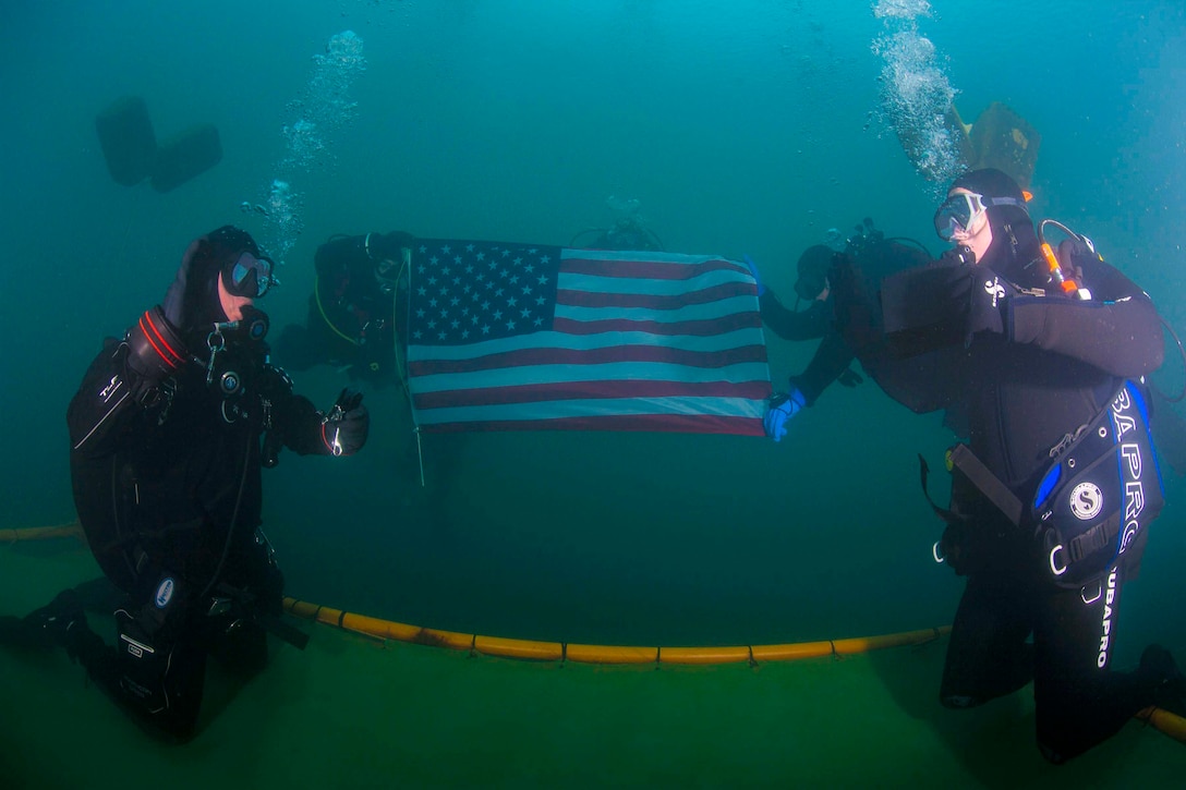 Two soldiers wearing diving gear face each other under water; two soldiers hold an American flag in the background.