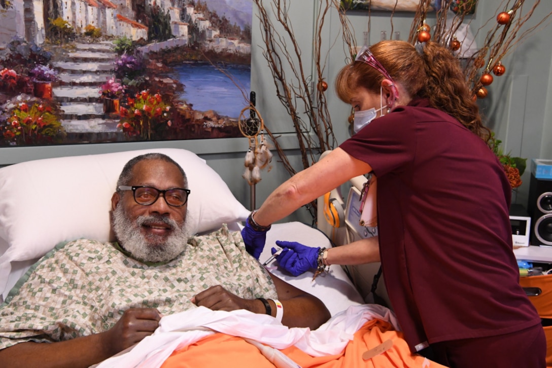 A man in a hospital bed wearing a hospital gown is injected in the left arm by a woman healthcare professional in dark red scrubs.