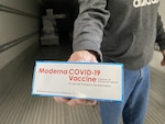 A box labeled "Moderna COVID-19 Vaccine" is held in a man's hand.