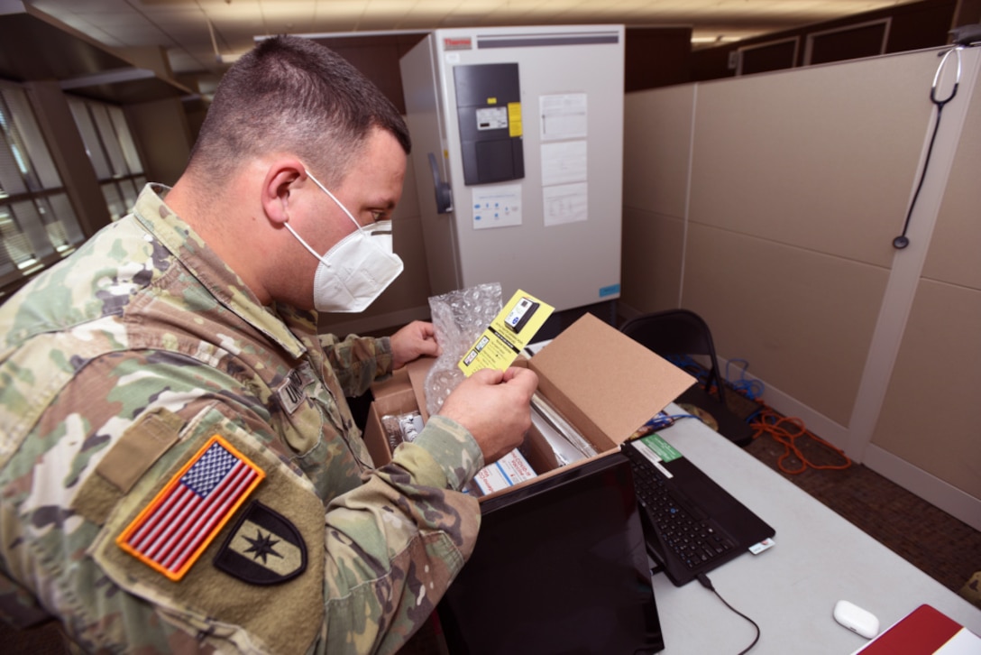 A man in operational camo pattern uniform and protective mask  unpackages a yellow card from a box.