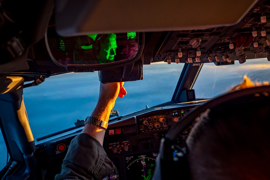 A Navy pilot's face is visible in a rear-view mirror in the cockpit of an aircraft he's flying over blue water.
