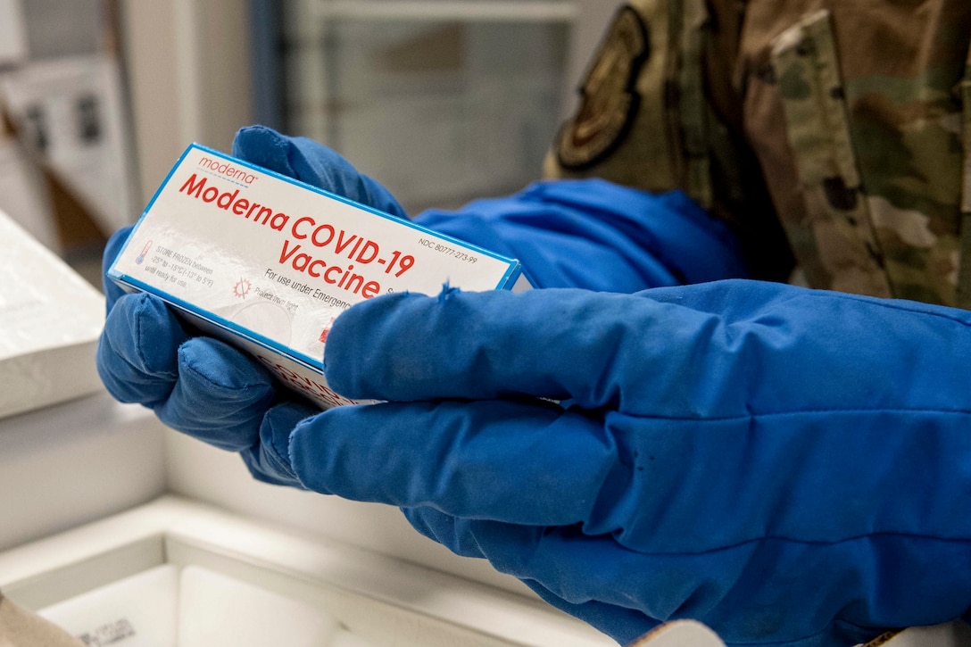 Blue gloves worn by an airman hold a box labelled "Moderna COVID-19 Vaccine."