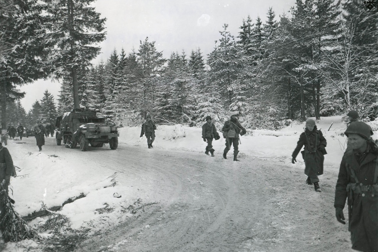 Soldiers march near a vehicle along a winding road in a snow-covered forest.