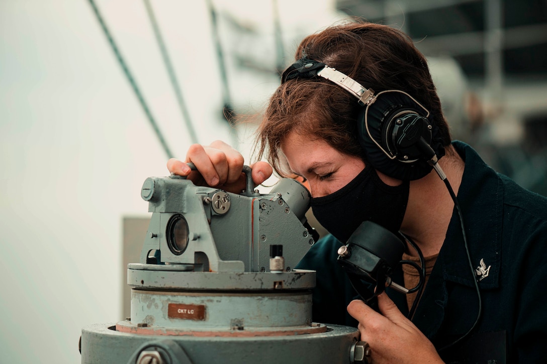 A sailor looks through a telescope while wearing headphones.