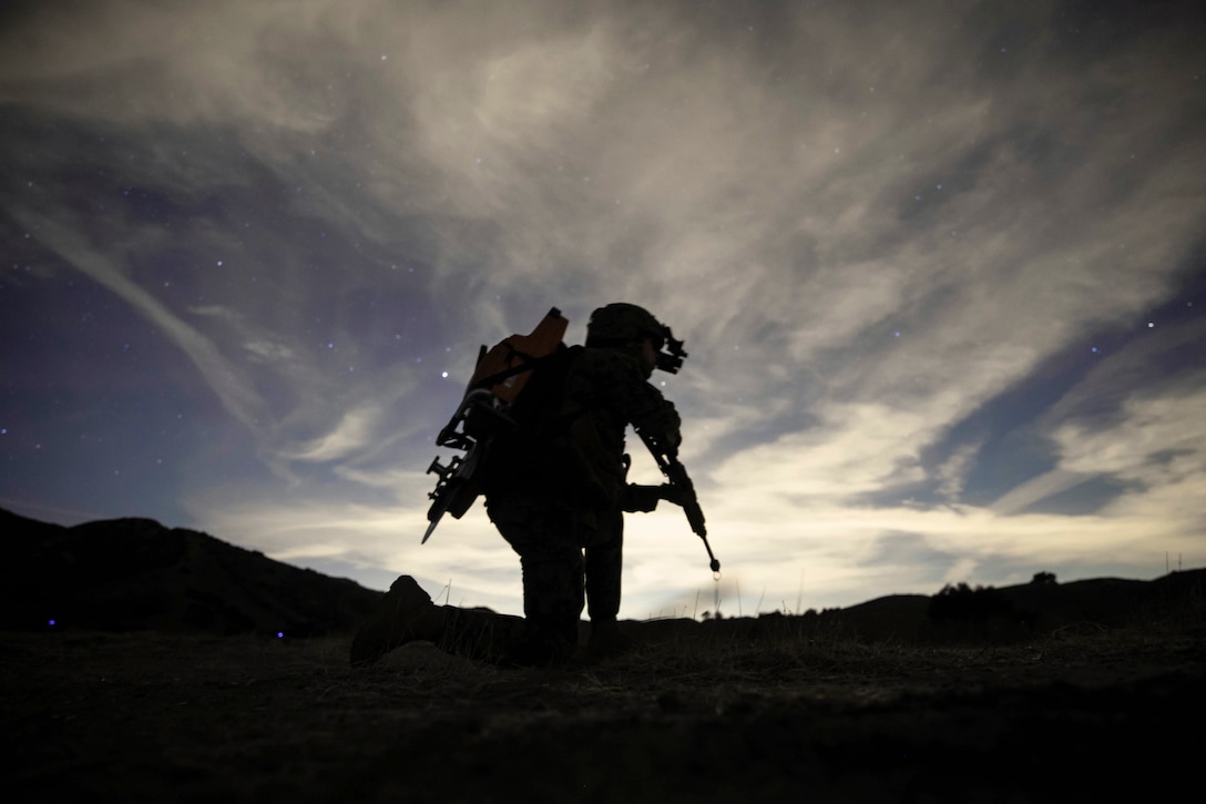 A Marine shown in silhouette holds a weapon while kneeling in a field.