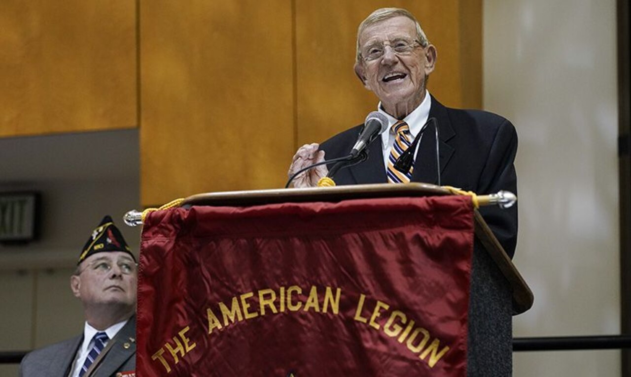 A man stands at a lectern and speaks into a microphone; the lectern is draped with a banner that reads "The American Legion."