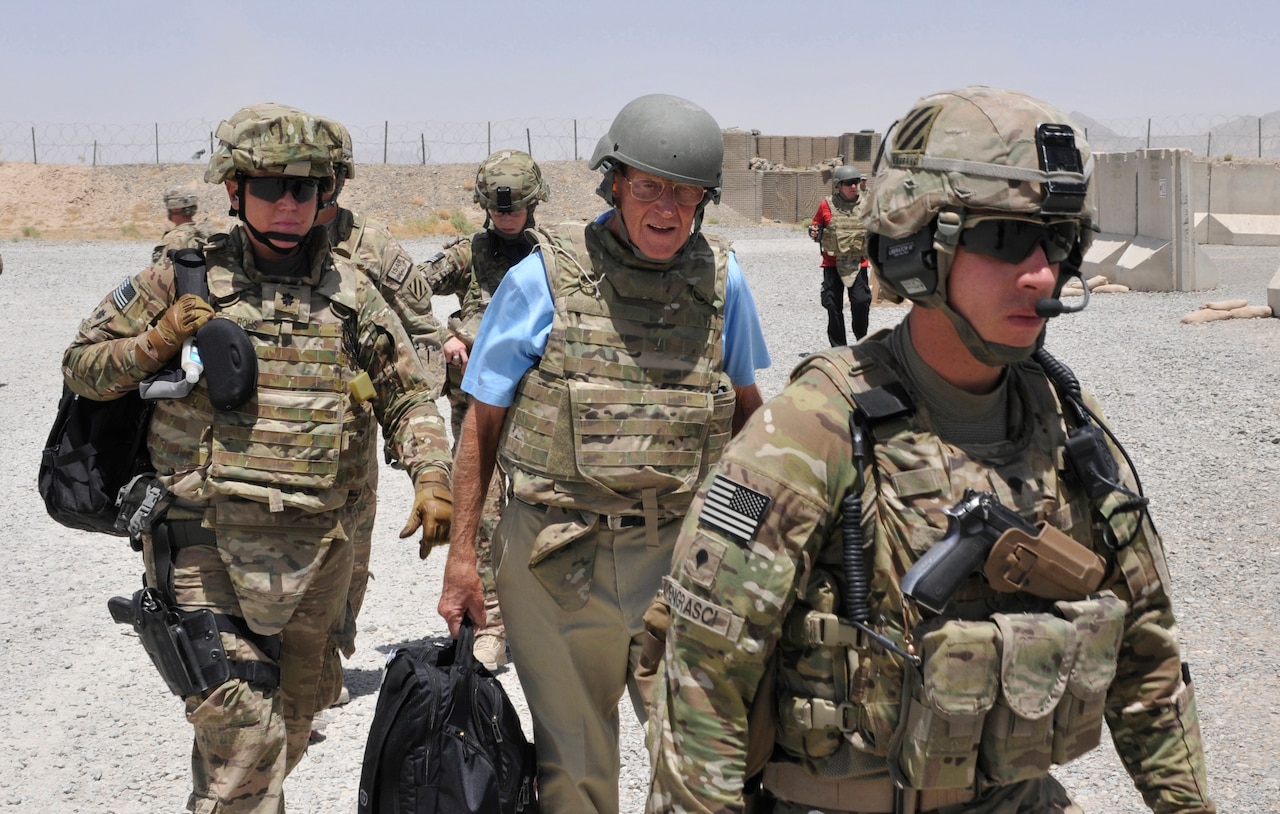 Three men dressed in combat gear walk together as service members stand behind them.