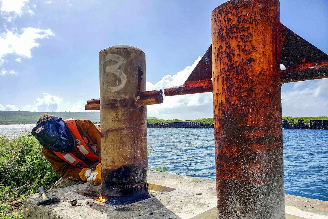 A sailor wearing a protective helmet uses a torch on a deteriorated mooring post.