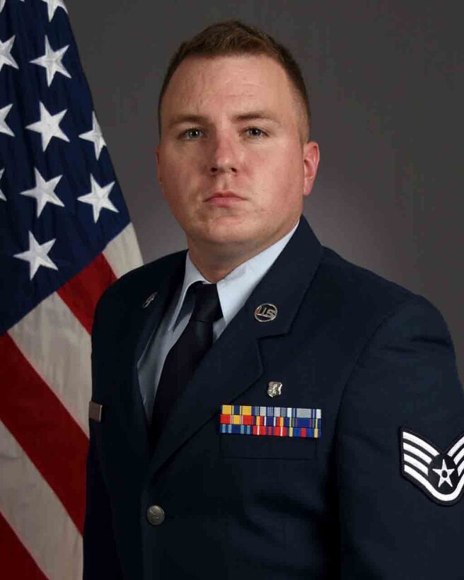 An airman in dress blues poses for a photo in front of a flag.