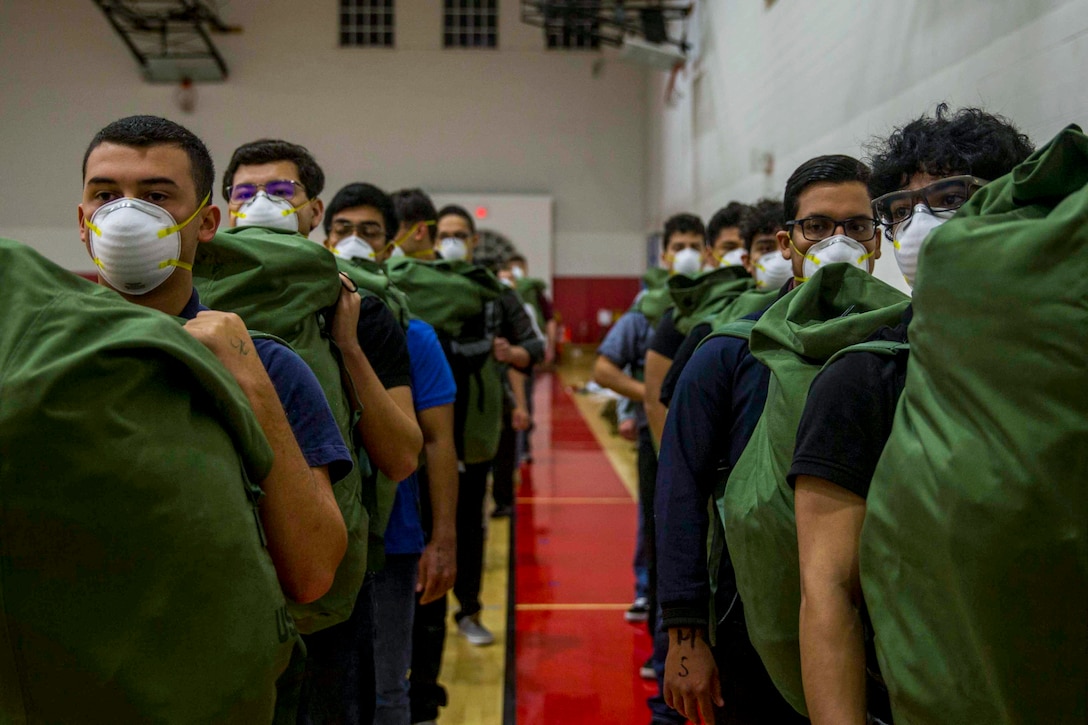 Marine Corps recruits wearing face masks and carrying sacks stand in two lines in a gym-type room.