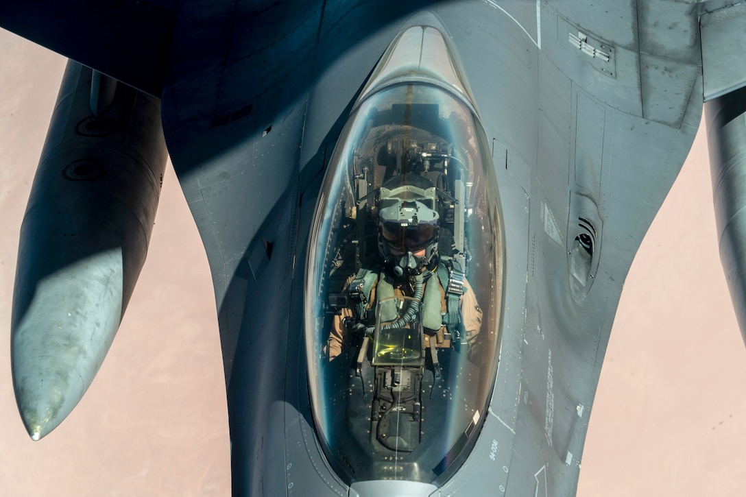 An airman, seen from overhead, sits in the cockpit of a jet flying over desert-type terrain.