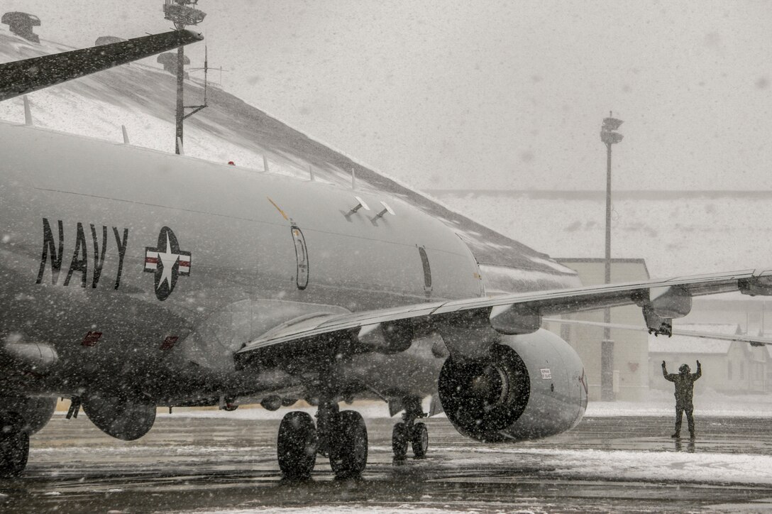A Navy aircraft sits on a snowy flightline as  a sailor signals in the background.