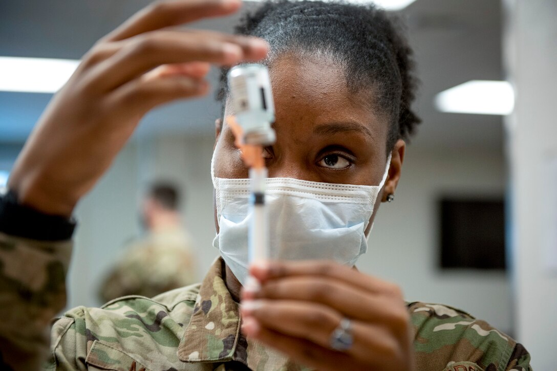 An airman holds a glass vial in front of her face and punctures it with a syringe.