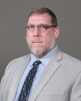 Headshot of a man with a short beard. He is wearing glasses, a light-gray suit, baby-blue shirt and dark tie.