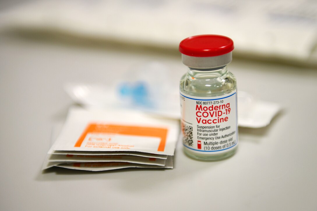 A vial sits on a whilte surface along with some packets of alcohol wipes.